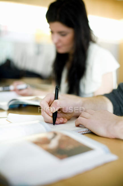 Vista cortada de mãos de homem escrevendo em livro com colega de classe feminina no fundo — Fotografia de Stock