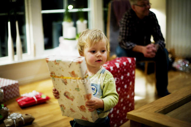 Niño pequeño sosteniendo regalo de Navidad con el hombre mayor en el fondo en casa - foto de stock