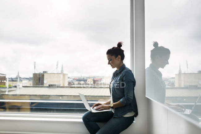 Seitenansicht der Frau mit Laptop auf Fensterbank im Büro — Stockfoto