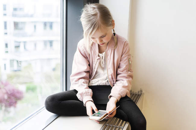 Девушка сидит на подоконнике со смартфоном — стоковое фото