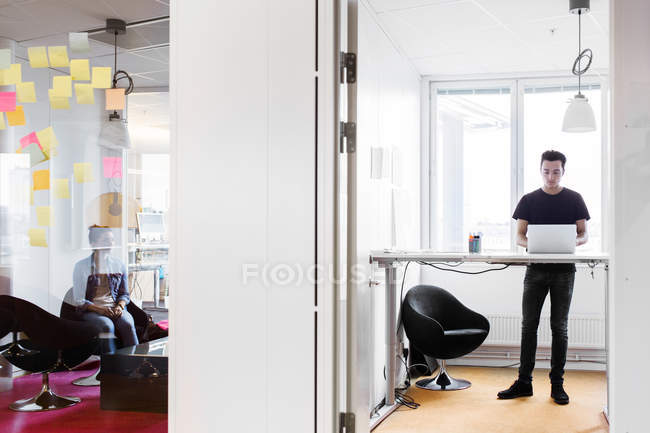 Homem de pé usando laptop no escritório com mulher sentada no quarto ao lado — Fotografia de Stock