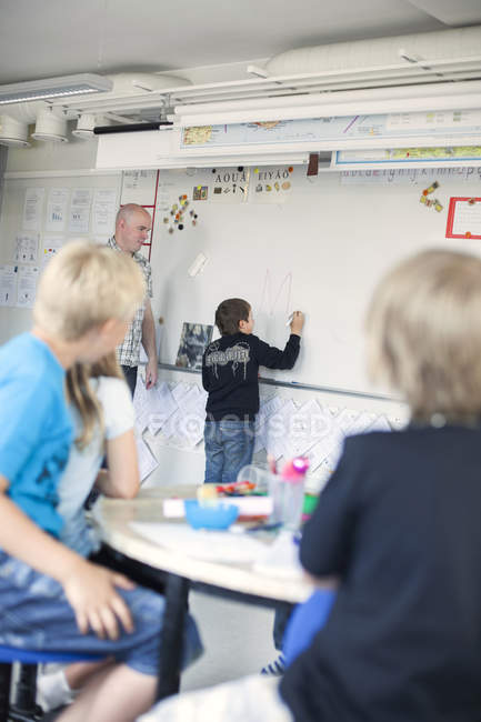 Professor olhando menino escrevendo em quadro branco em sala de aula — Fotografia de Stock