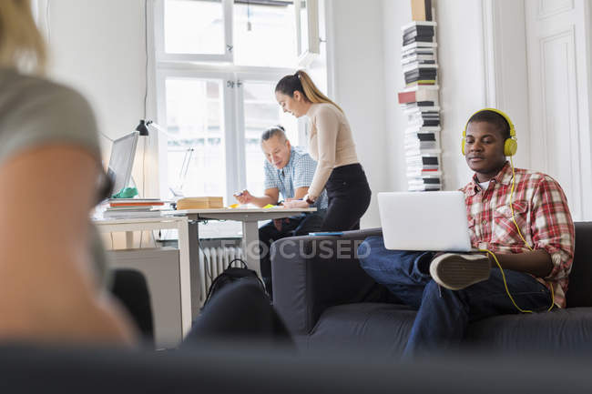 Работник с помощью ноутбука на диване и прослушивания музыки, коллеги, работающие в фоновом режиме — стоковое фото