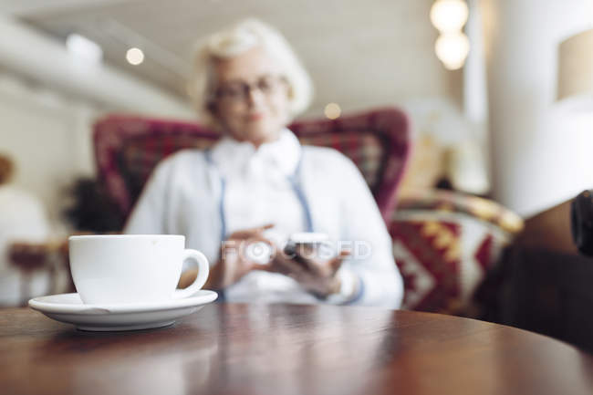 Coffee cup on table and senior woman using mobile phone during coffee break in cafe — Stock Photo