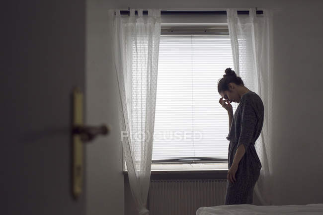 Mujer joven pensativa de pie junto a la ventana - foto de stock