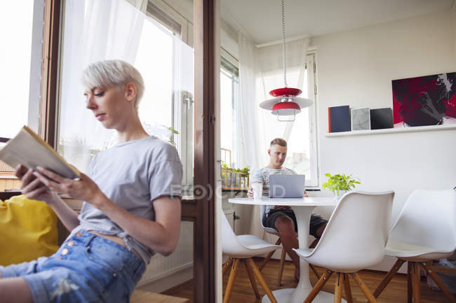 Woman reading book on balcony, man using laptop in dining room — Stock Photo