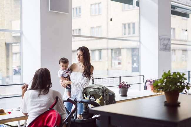 Mother holding baby son (6-11 months) and talking with friend in cafe — Stock Photo