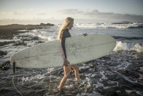 Femme avec planche de surf dans les mains marchant sur les rochers — Photo de stock