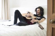 Чоловік лежить на ліжку і грає на гітарі — стокове фото