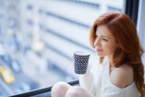 Donna con bevanda calda seduta vicino alla finestra — Foto stock