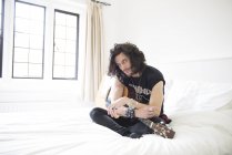 Junger Mann sitzt mit Gitarre im Bett — Stockfoto