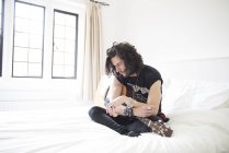 Молодой человек, сидящий на кровати с гитарой — стоковое фото