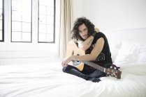 Молодой человек, сидящий на кровати с гитарой — стоковое фото