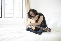 Junger Mann sitzt mit Gitarre im Bett — Stockfoto