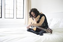 Joven sentado en la cama con la guitarra - foto de stock