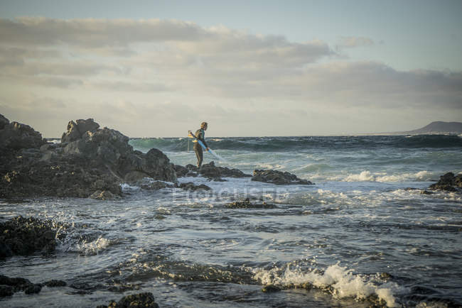 Человек с доской для серфинга в руках, шагающий по скалам — стоковое фото