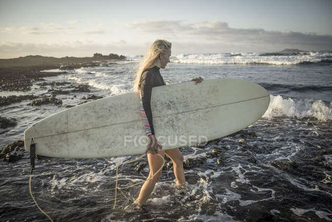 Женщина с доской для серфинга в руках ходит по камням — стоковое фото