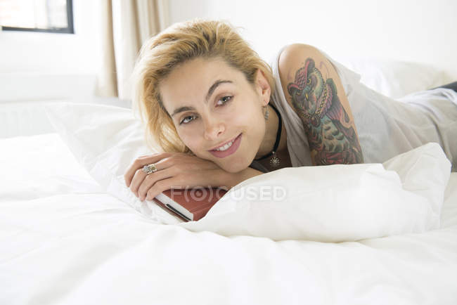 Жінка з татуюваннями лежить на ліжку з книгою — стокове фото
