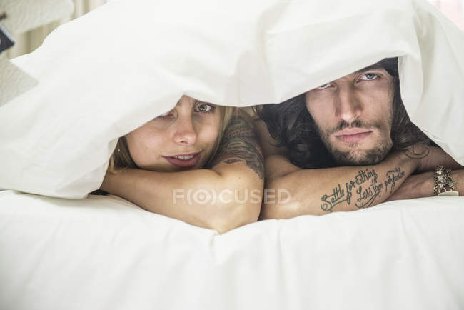 Tätowiertes Paar guckt unter Bettdecke hervor — Stockfoto