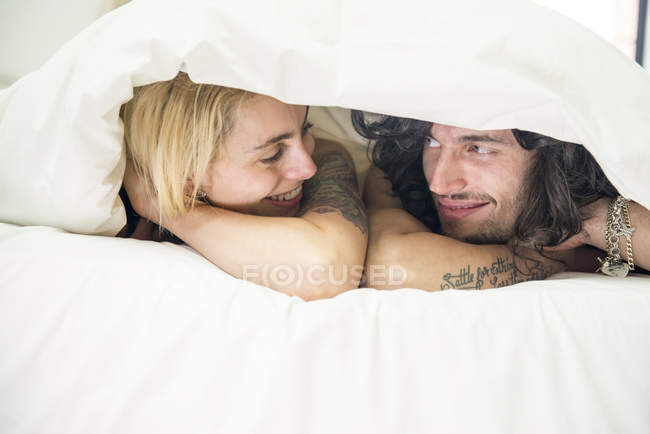Tätowiertes Paar guckt unter Bettdecke hervor — Stockfoto