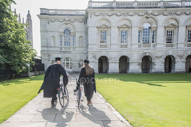 Étudiants en robes de remise des diplômes poussant des vélos — Photo de stock