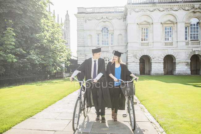 Студенты в выпускных платьях толкают велосипеды — стоковое фото