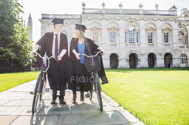 Estudantes em vestidos de graduação empurrando bicicletas — Fotografia de Stock