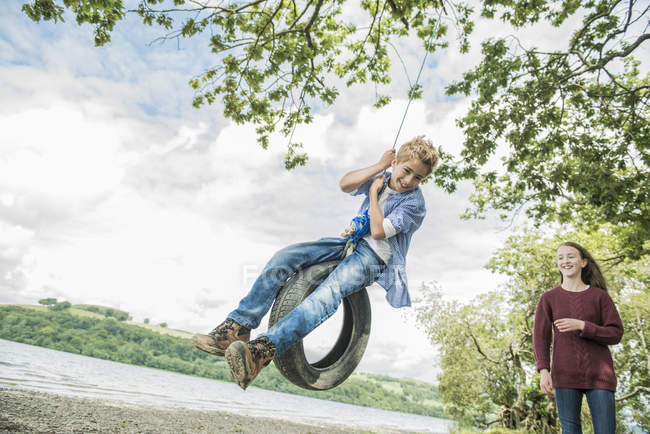 Девочка и мальчик играют на висящей на дереве шине — стоковое фото
