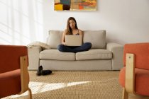 Молодая женщина сидит скрестив ноги на диване, используя ноутбук — стоковое фото