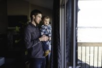 Hombre e hijo pequeño mirando por la ventana en casa - foto de stock