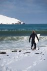 Surfista che porta la tavola da surf sulla spiaggia innevata — Foto stock