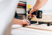Cropped view of young man in carpentry workshop using cordless screwdriver, screwing into plywood — Stock Photo
