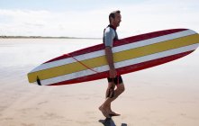 Surfeur mâle portant la planche sur la plage — Photo de stock