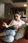 Mujer leyendo libro en sofá en casa - foto de stock