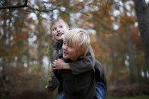 Boy giving friend piggyback through autumnal forest — Stock Photo