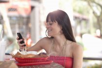 Молода жінка тримає мобільний телефон у відкритому кафе, посміхаючись — стокове фото