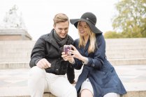 Young couple sitting on step reading smartphone texts, London, England, UK — Stock Photo