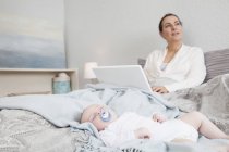 Mère utilisant un ordinateur portable pendant que le bébé garçon dort à côté — Photo de stock