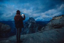 Young woman taking photograph on rock overlooking Yosemite National Park at dusk, California, USA — Stock Photo