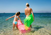 Mulher com a menina que vai nadar na água do mar e carregando o colchão inflável, vista traseira — Fotografia de Stock