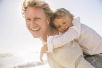 Батько на пляжі дає синові свинарство, дивлячись на камеру посміхаючись — стокове фото