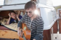 Середній дорослий чоловік грає на гітарі позаду кемперського фургона, посміхаючись — стокове фото