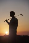 Golfista llevando palo de golf sobre el hombro delante de la puesta del sol, mirando hacia otro lado - foto de stock