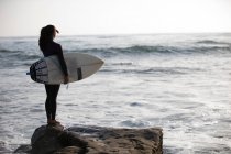Молодая женщина, стоящая на скалах с доской для серфинга — стоковое фото