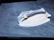 Vista en ángulo alto del filete de lenguado de limón crudo y la cuña de limón en papel de hornear y pizarra - foto de stock