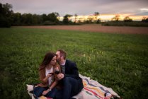 Romantic couple with red wine relaxing on picnic blanket in field at sunset — Stock Photo