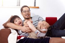 Молодая, современная китайская семья отца и маленького сына, сидящих на диване и вместе смотрящих телевизор дома — стоковое фото
