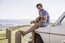 Mid adult man sitting on car at coast playing guitar, Cape Town, South Africa — Stock Photo