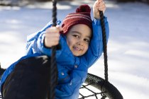 Niño preescolar con sombrero de punto en hamaca swing, mirando a la cámara sonriendo - foto de stock