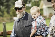 Бородатий дід у бейсбольній кепці та сонцезахисних окулярах, що носять онука — стокове фото